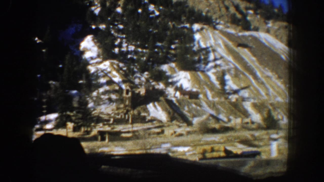 8mm home movie frame showing a steep striated cliff face with snow, conifers, and structures at its base, shot from a moving car in the Aspen, Colorado area, 1961.