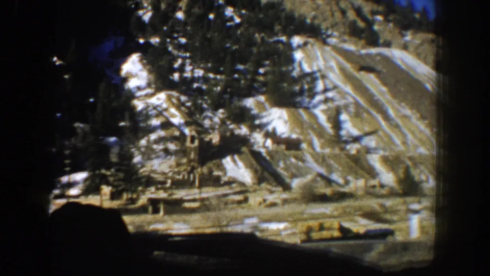 8mm home movie frame showing a steep striated cliff face with snow, conifers, and structures at its base, shot from a moving car in the Aspen, Colorado area, 1961.