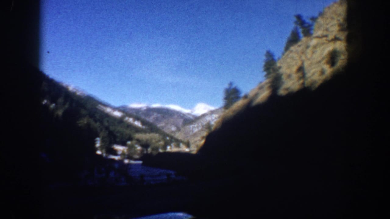 8mm footage of a broad Colorado mountain valley with snow-dusted peaks, conifers, deep blue sky, and a small valley settlement, shot from a moving car circa 1961.