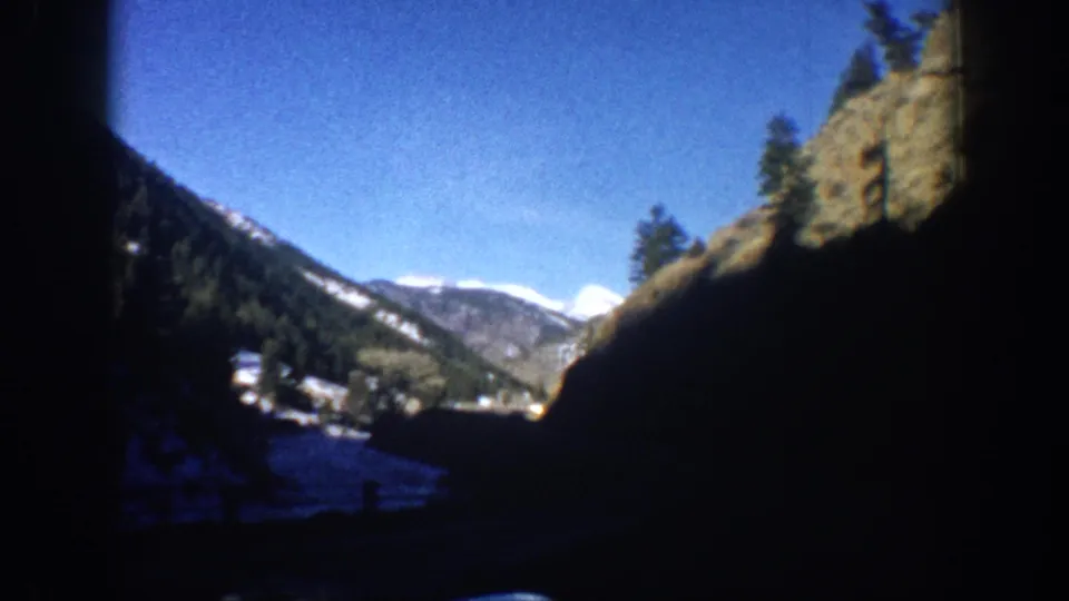 8mm POV shot from a moving car showing steep canyon walls, conifer forest, snow on lower slopes, and a valley settlement in the Aspen, Colorado area, circa 1961.