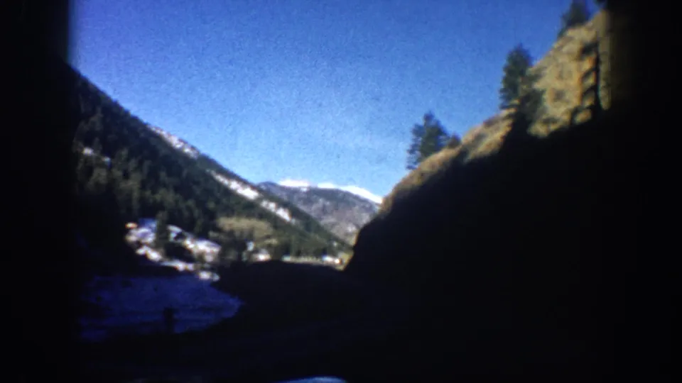8mm home movie frame of a Colorado mountain valley with rocky slopes, evergreen trees, snow patches, and a small settlement filmed from a moving car, circa 1961.