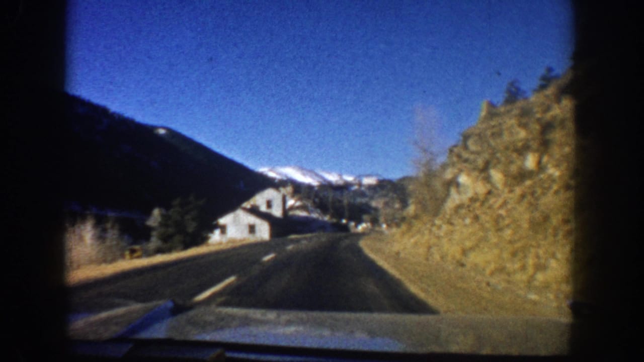 POV driving shot approaching a small Colorado mountain town, circa 1961, with snow-capped peaks, rocky outcroppings, white painted structures, and clear blue sky.
