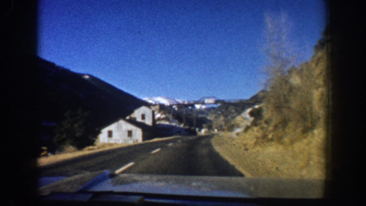 Continuing windshield driving shot: two-lane road curves toward white painted structures, rocky cliff on the right, snow-capped peaks visible in the background.