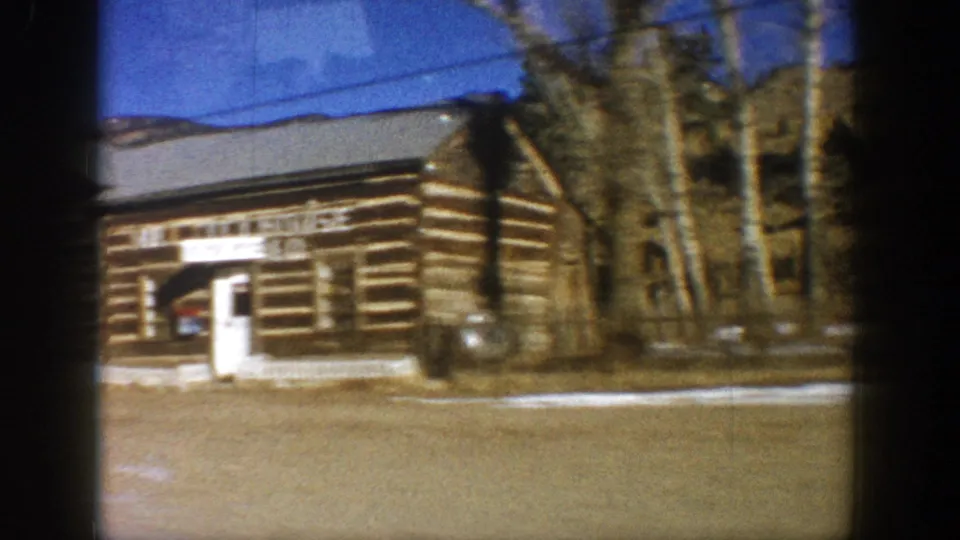 Large log commercial building with partially visible signage seen from a moving vehicle in a Rocky Mountain town, circa 1961; bare trees and snow patches visible.