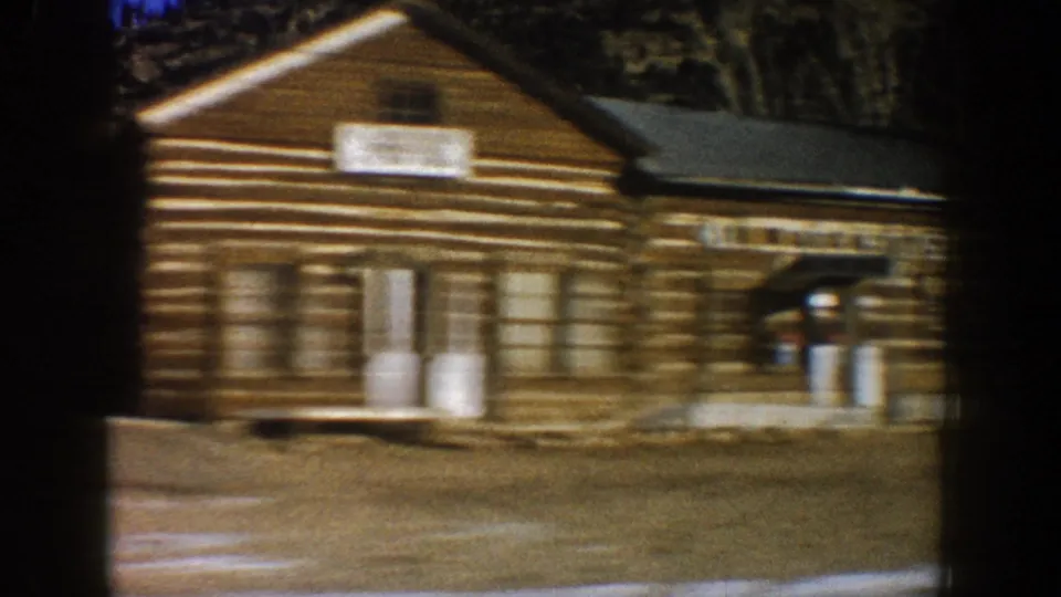 Large log building with motion-blurred signs viewed from a moving car in a Rocky Mountain town, circa 1961; utility poles and snow patches visible.