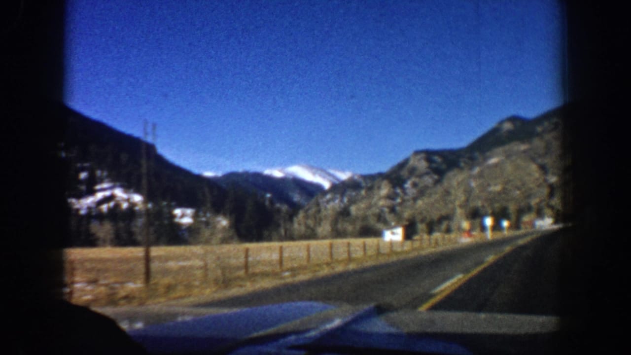 8mm home-movie windshield POV on a two-lane road near Aspen, Colorado, circa 1961, with snow-capped Rocky Mountains ahead.