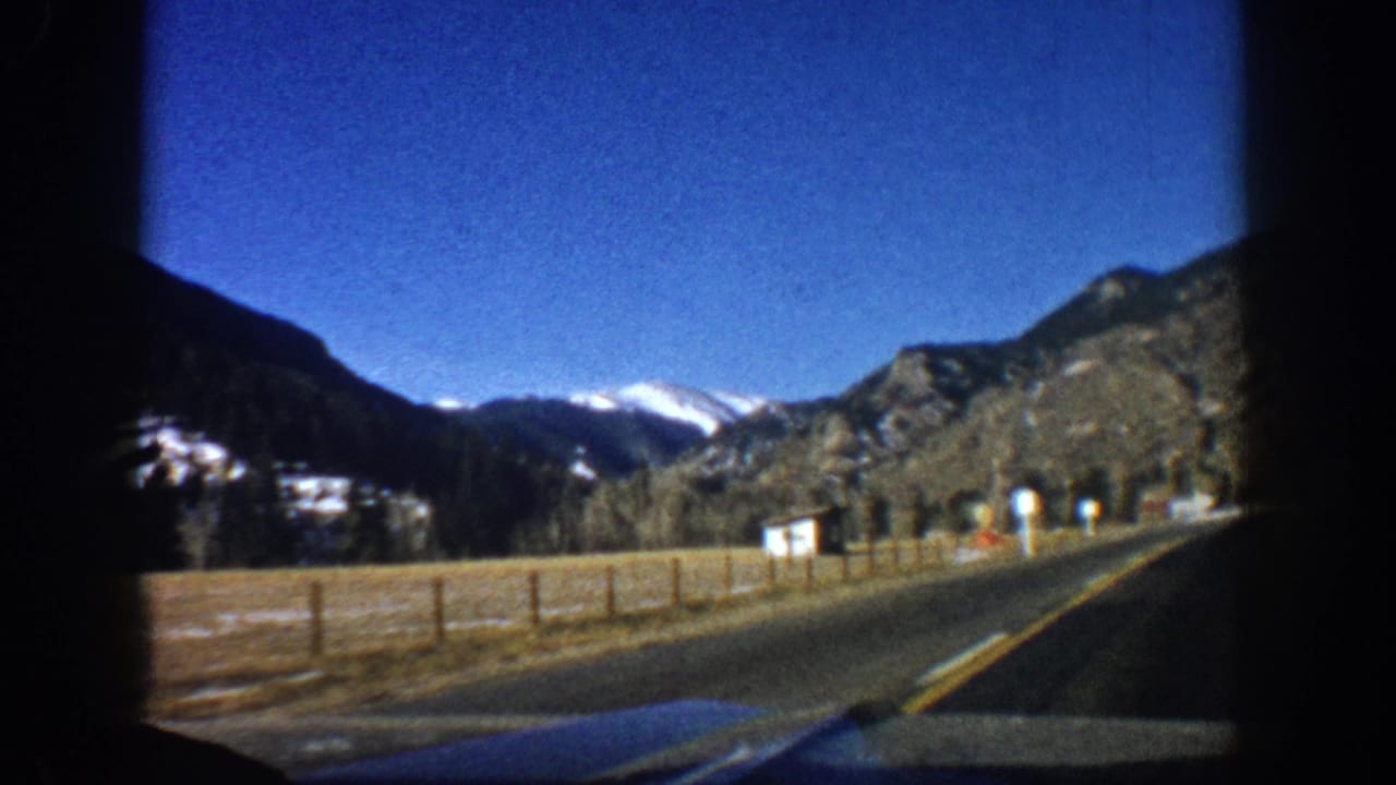 8mm windshield POV on two-lane road approaching Aspen, Colorado, circa 1961, with snow-capped peaks, wooden fencing, and roadside markers.