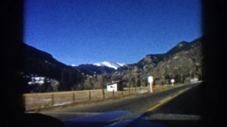 8mm driving POV on two-lane road near Aspen, Colorado, circa 1961, with bare trees, wooden fence, signpost, and mountain backdrop.
