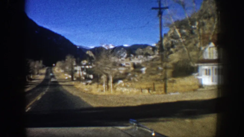 8mm driving POV approaching a small settlement near Aspen, Colorado, circa 1961; white building, utility pole, and Rocky Mountain backdrop visible.