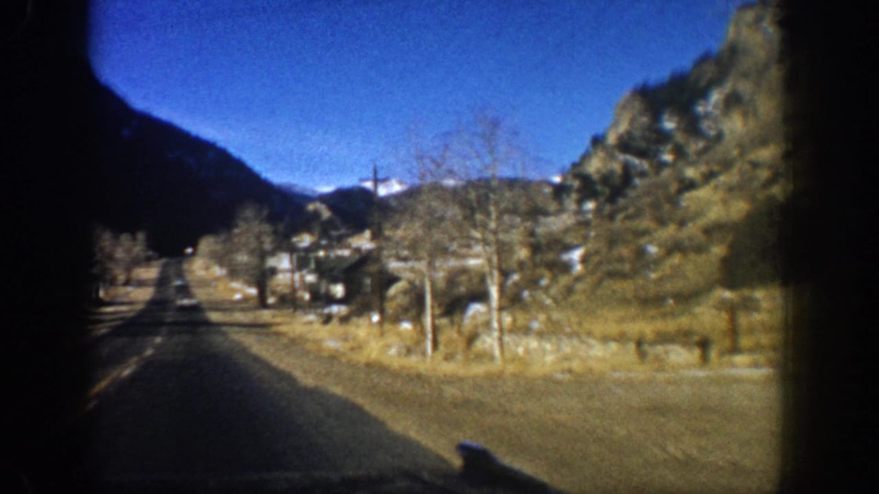 Two-lane mountain road through a valley with bare trees, rocky peaks, and deep blue sky, shot from inside a moving vehicle.