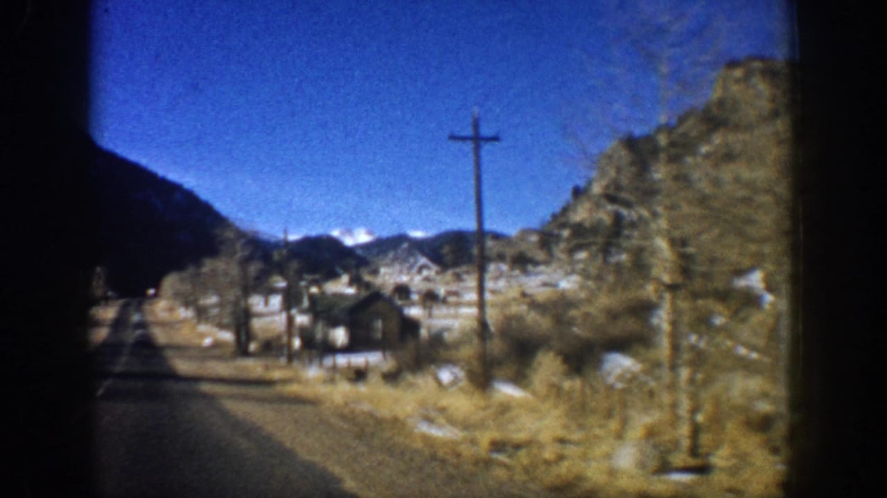 Moving-vehicle POV on a Colorado mountain road, circa 1961; wooden utility pole, rocky hillside, and small roadside cabins visible alongside the road.