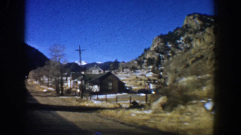 A dark-timbered cabin with a fence beside a Colorado mountain road, circa 1961; snow patches on the ground and a rocky escarpment visible behind the structure.