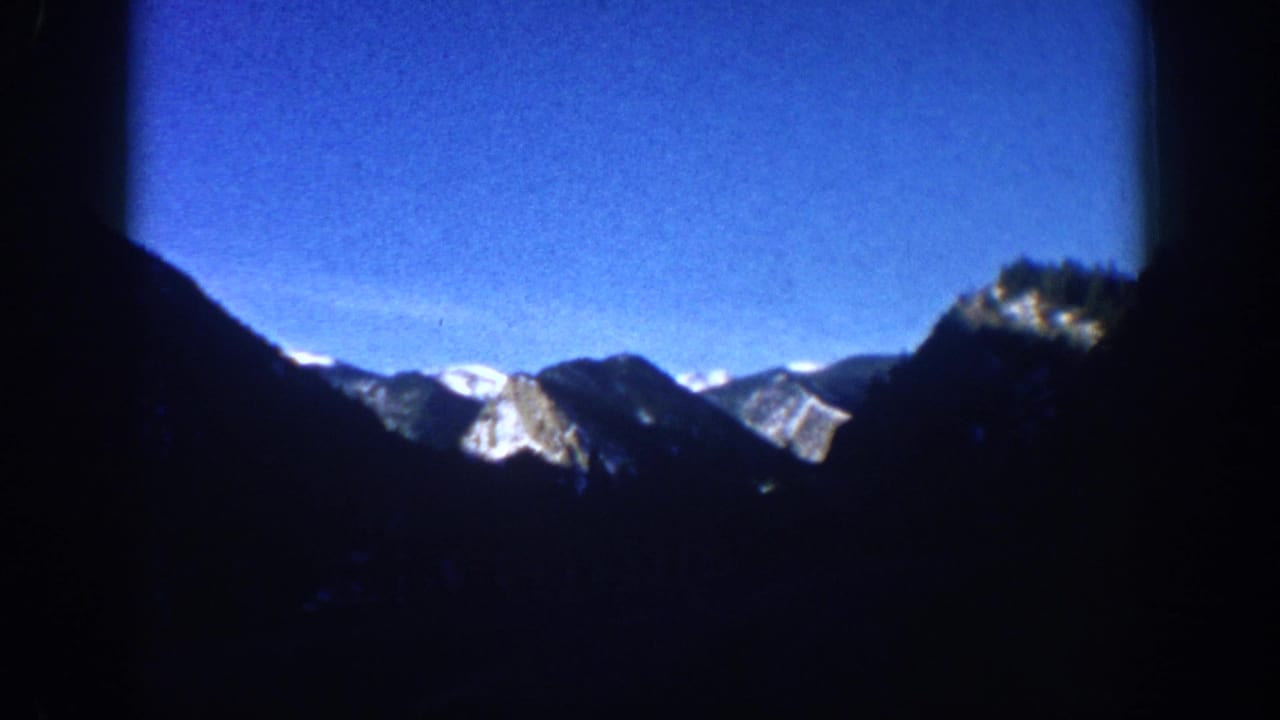 8mm home movie frame circa 1961, wide establishing view of snow-capped Rocky Mountains near Aspen, Colorado, with silhouetted valley foreground and brilliant blue sky.