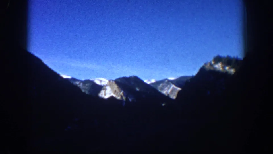 8mm home movie frame circa 1961, wide establishing view of snow-capped Rocky Mountains near Aspen, Colorado, with silhouetted valley foreground and brilliant blue sky.