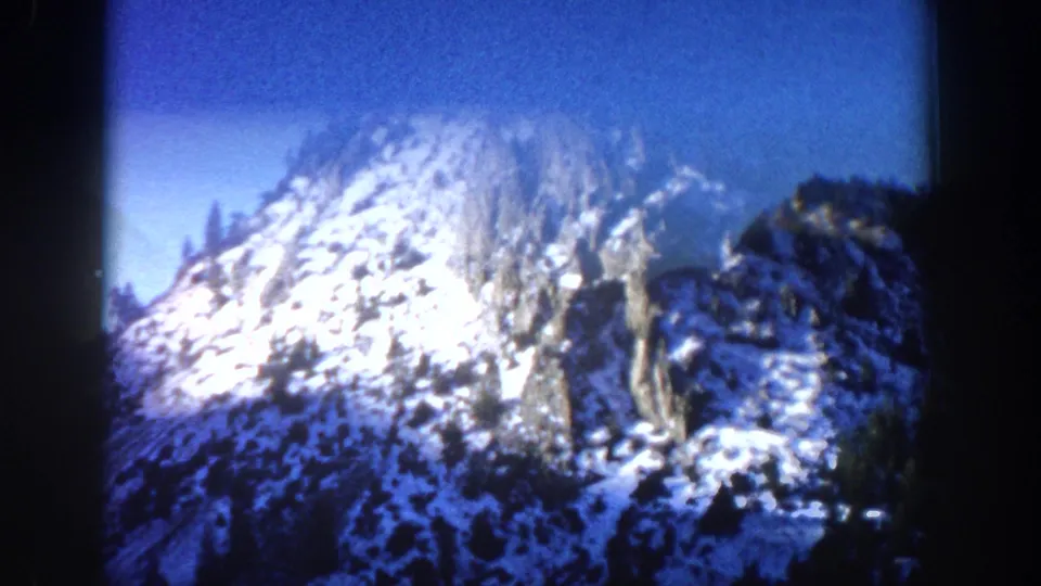 8mm home movie frame circa 1961, close-up of a sunlit snow-covered rocky peak near Aspen, Colorado, with sparse conifers at lower edges and cobalt blue sky.
