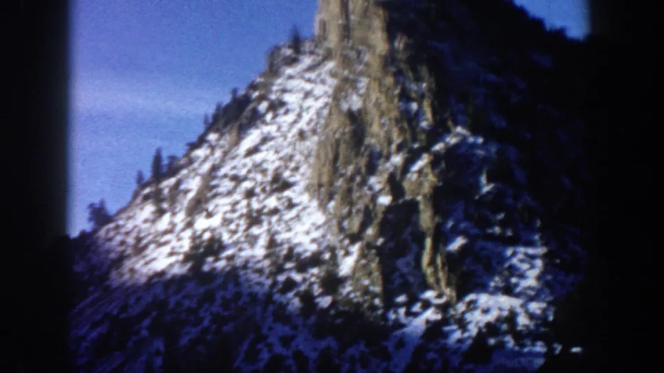 8mm home movie frame circa 1961, upward-angle view of a dramatic rocky spire with patchy snow and sparse conifers, likely in the Aspen, Colorado area.