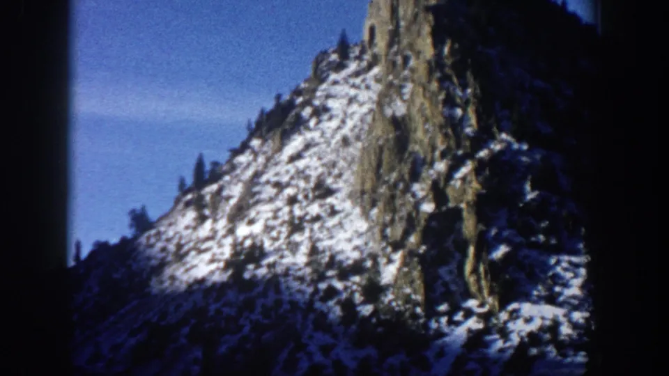 8mm home movie frame circa 1961, upward view of a snow-dusted rocky pinnacle near Aspen, Colorado, with evergreen trees on the slope and a clear blue sky.