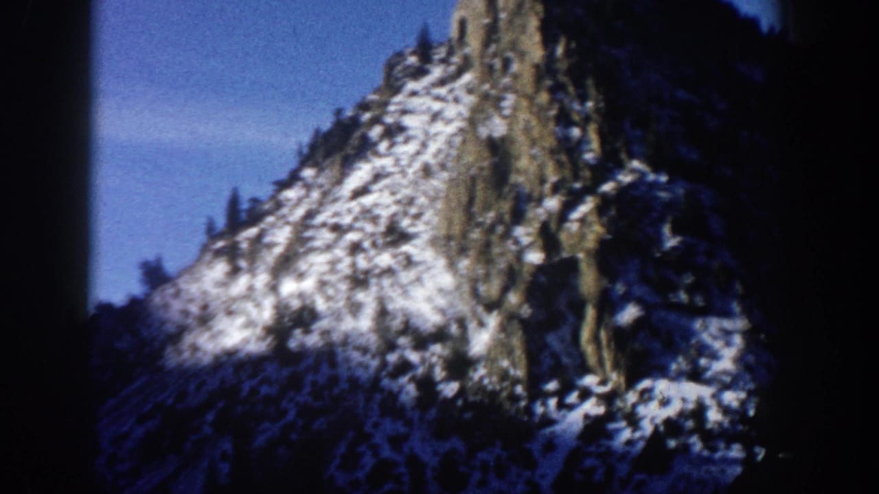 1961 8mm home movie frame, Aspen Colorado area: snow-patched rocky spire framed at a steep upward angle with heavy circular vignetting and bare trees at the crest.
