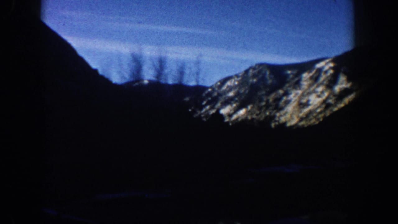 1961 8mm home movie frame, Aspen Colorado area: mountain ridge silhouetted against afternoon blue sky with thin contrail or cloud streaks and a snow-patched distant slope.