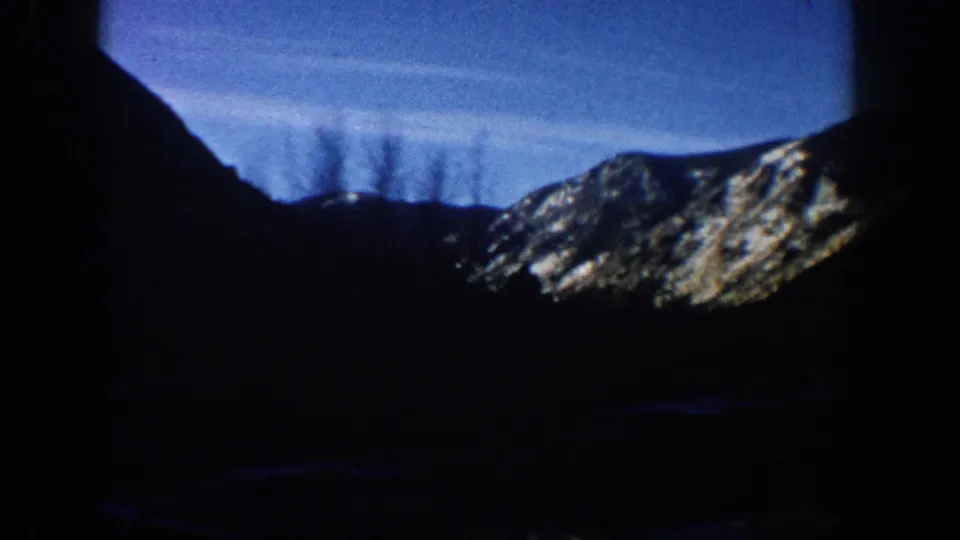 1961 8mm home movie frame, Aspen Colorado area: mountain ridge silhouetted against afternoon blue sky with thin contrail or cloud streaks and a snow-patched distant slope.