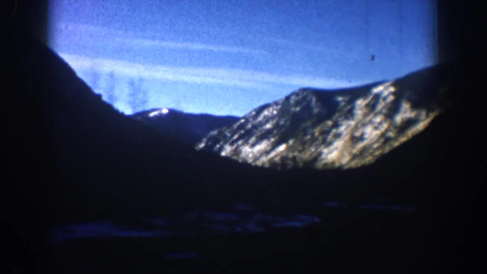 1961 8mm home movie frame, Aspen Colorado area: wide alpine valley panorama showing snow-patched mountain flanks under a bright blue sky with contrail streaks.