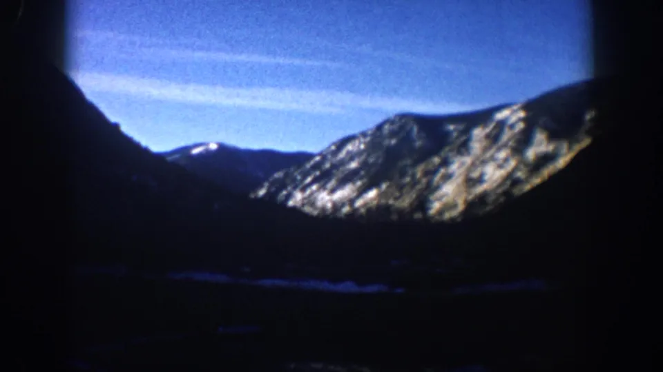 1961 8mm home movie pan frame, Aspen Colorado area: snow-patched alpine slopes under blue sky, part of a slow camera pan across the mountain range.