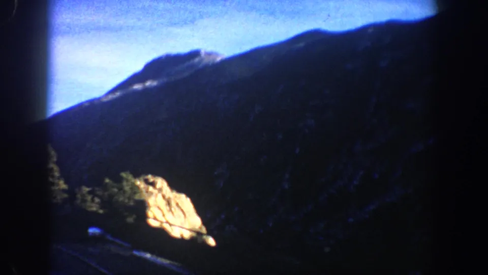 Sunlit rock outcrop and dark evergreen conifers framing mountain ridge under blue sky, Colorado Rockies circa 1961, 8mm film.