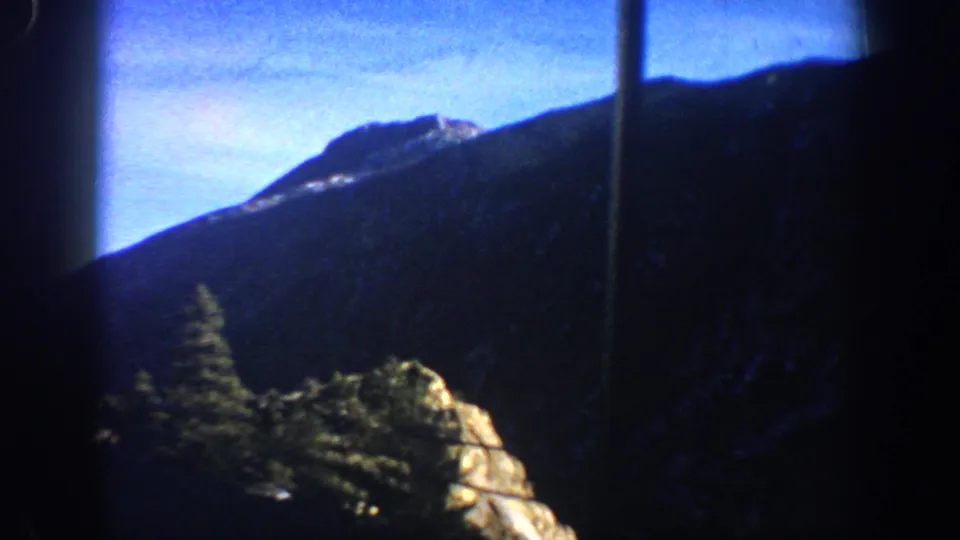 Dark evergreen conifers and rocky outcrop beneath mountain ridge and blue sky, Colorado Rockies circa 1961, 8mm film.