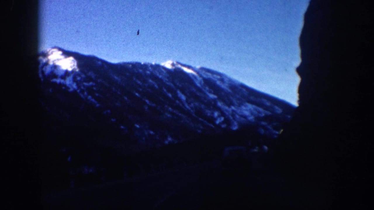 Snow-capped mountain peak against deep blue sky with dark rocky ridge silhouette on right, 8mm film