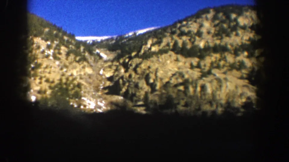 Sunlit rocky mountain face densely covered with conifers and snow patches, Aspen Colorado area, 1961, 8mm film