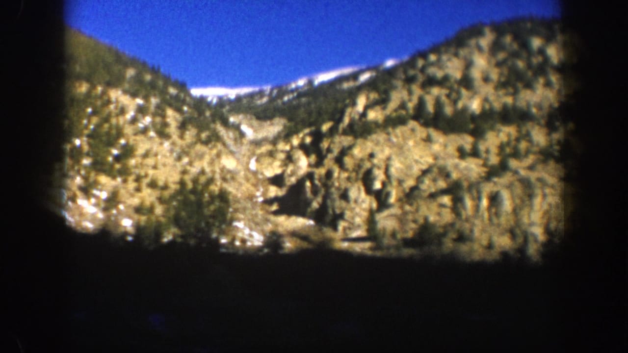 8mm home movie frame from Aspen, Colorado, 1961, showing a sunlit rocky mountain slope with snow-capped ridge and evergreen trees against a blue sky.