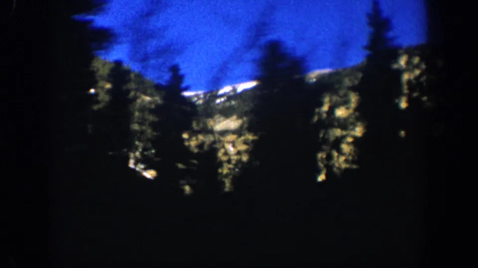 Silhouetted conifers frame a snow-capped Rocky Mountain ridge under a deep blue sky in this 1961 8mm frame from the Aspen, Colorado area.
