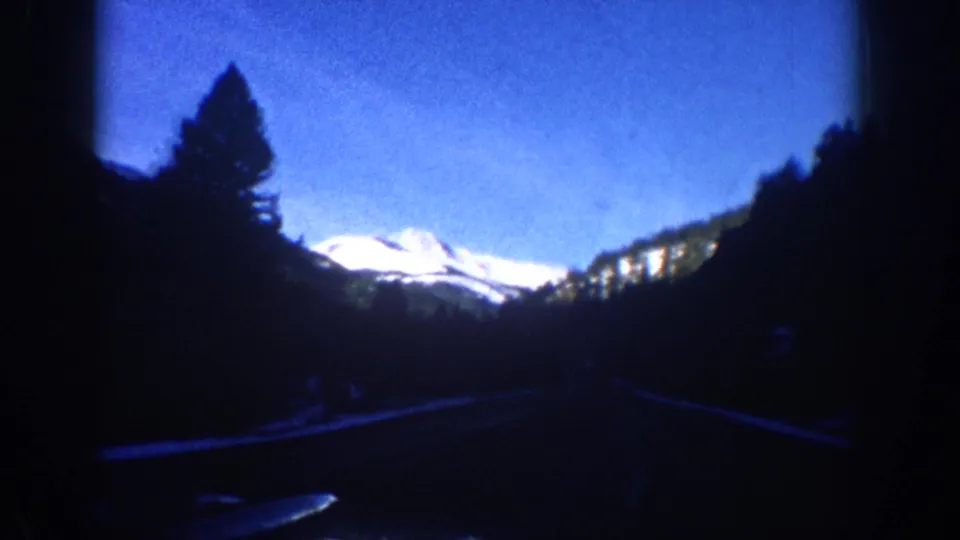 Roadside 8mm frame from the Aspen, Colorado area, 1961, showing a snow-covered mountain peak rising above silhouetted conifers with a road surface in the foreground.