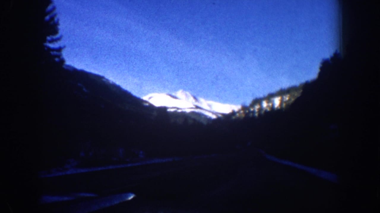 8mm home movie frame from what may be the Aspen, Colorado area circa 1961 — dark foreground treeline and a faint road edge below a bright snow-capped peak.