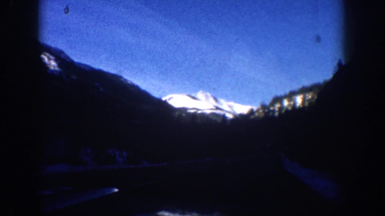 8mm frame from what may be Aspen, Colorado circa 1961 — same snow-capped peak from a slightly shifted vehicle position, with a dust-spot artifact near the top center.