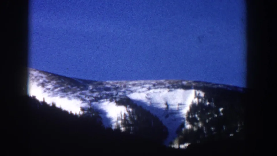 8mm home movie frame from what may be Aspen, Colorado circa 1961 — camera has panned to reveal a broader snow-covered ridge under a wide blue sky with tree silhouettes at the edges.