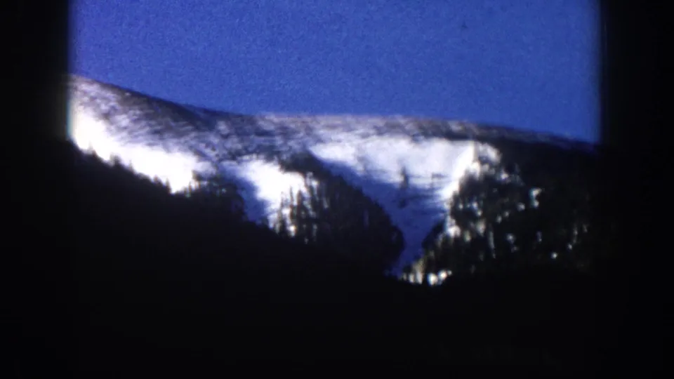 8mm frame from what may be Aspen, Colorado circa 1961 — a well-exposed snow slope dominates the right of frame with dark conifer silhouettes below and residual blue sky above.