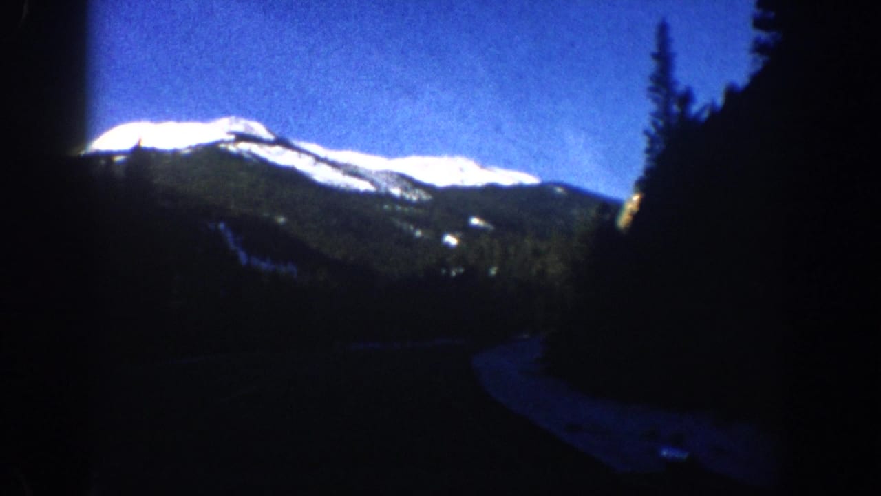 Prominent snow-capped mountain peak centered against blue sky with conifer trees at right edge, 8mm vignette