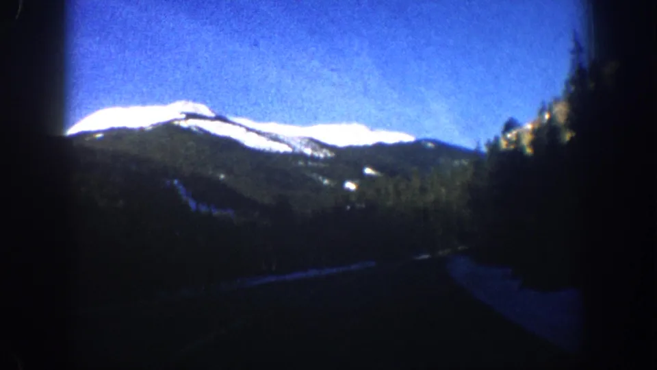 8mm home movie frame circa 1961 showing a Colorado mountain range from a moving vehicle, road surface appearing at the lower edge