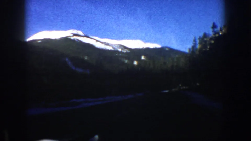 8mm home movie frame circa 1961 showing Colorado Rockies scenery from a moving vehicle, road visible at lower edge with characteristic blue color cast