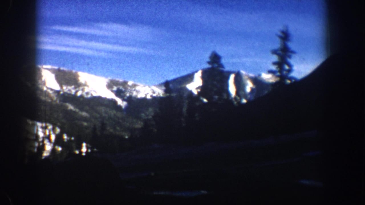 8mm home-movie frame from circa 1961 showing sunlit snow-capped mountain peaks and conifer silhouettes through a vehicle window, with a characteristic blue color cast.
