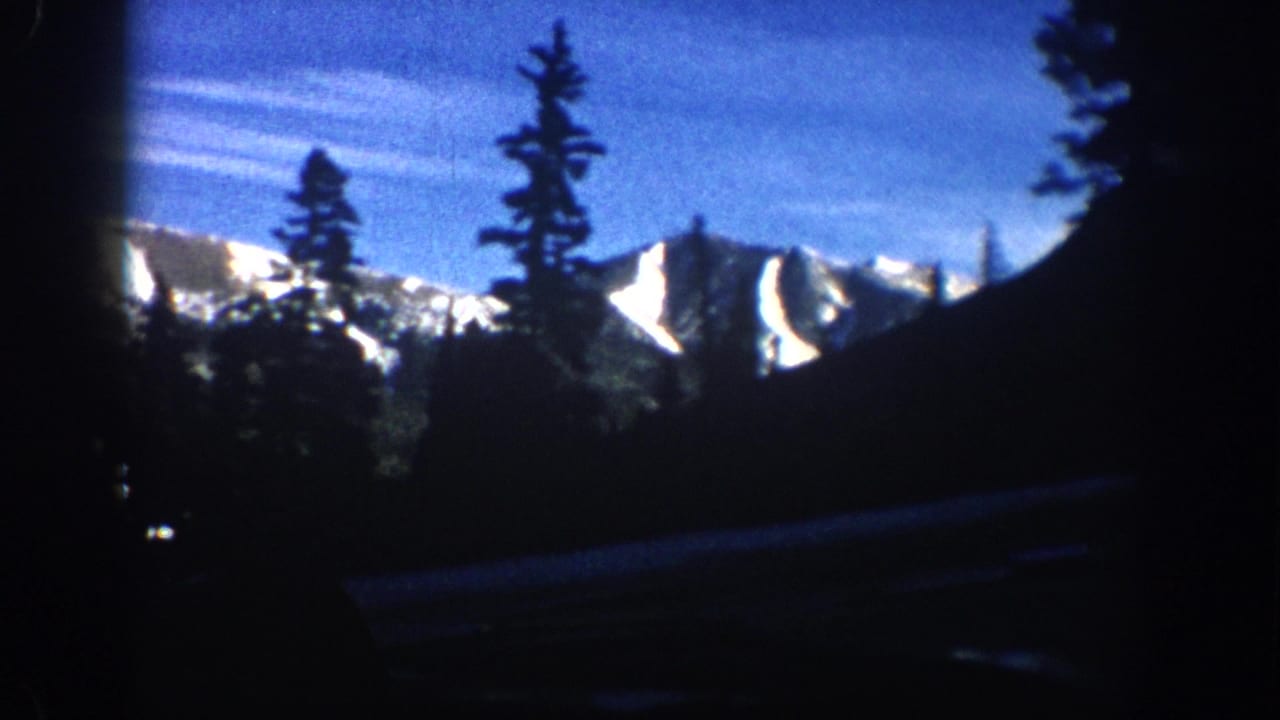 Vehicle-window view of a snow-covered mountain ridge and conifer trees against a deep blue sky, 8mm film with visible grain.