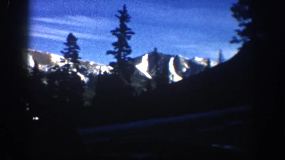 8mm footage from circa 1961 showing a slightly shifted angle of the same sunlit mountain ridgeline and conifer silhouettes, retaining the blue color cast of aged reversal film.