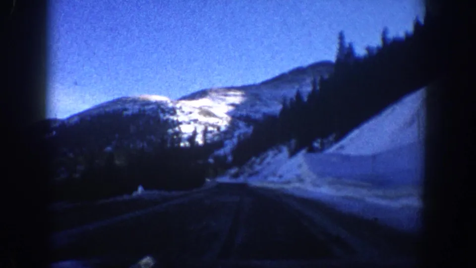 8mm home-movie frame from circa 1961 showing a snow-packed mountain road with a prominent snow-covered peak ahead, consistent with a Colorado Rocky Mountain drive.