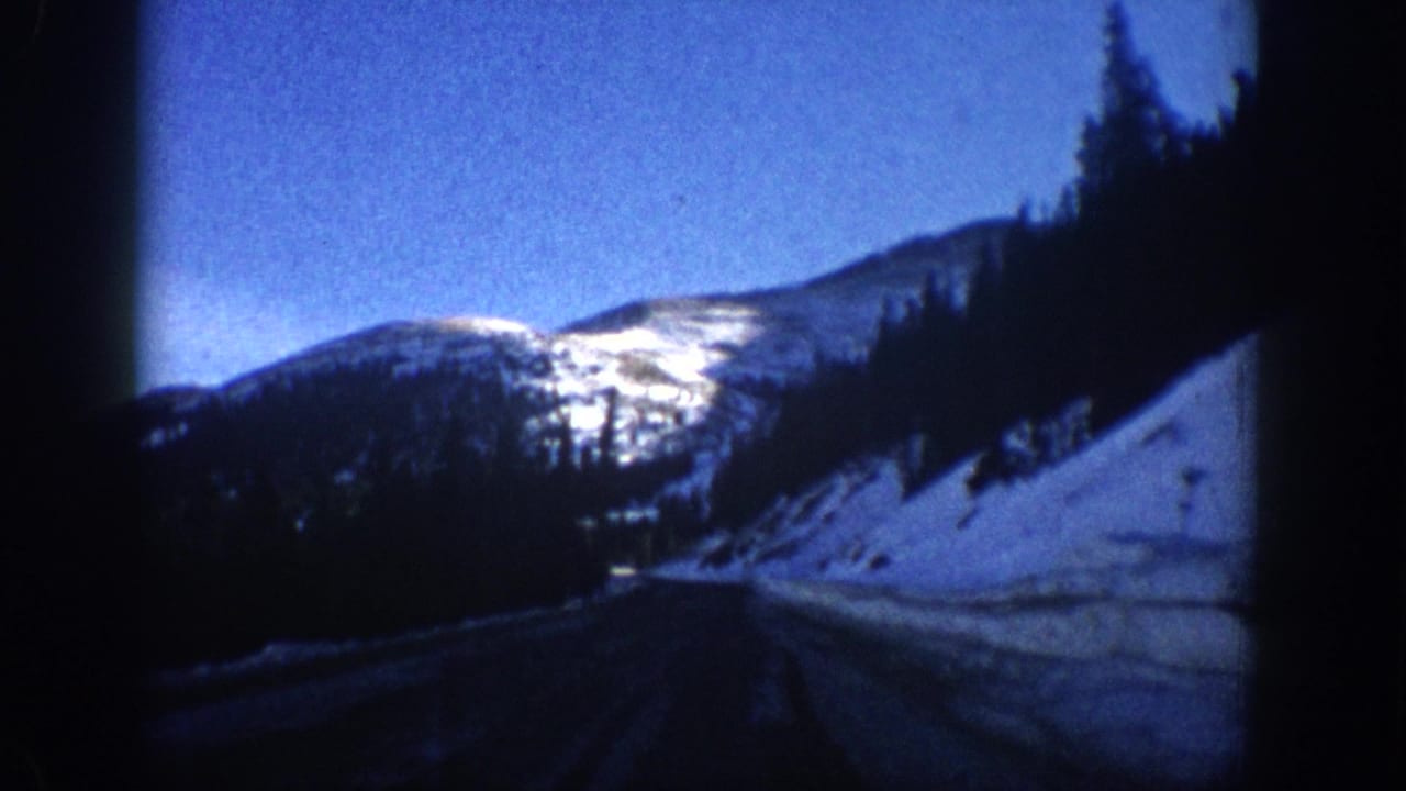 8mm home-movie frame showing a winding mountain road flanked by snow and conifers, likely Colorado Rockies area, circa 1961.