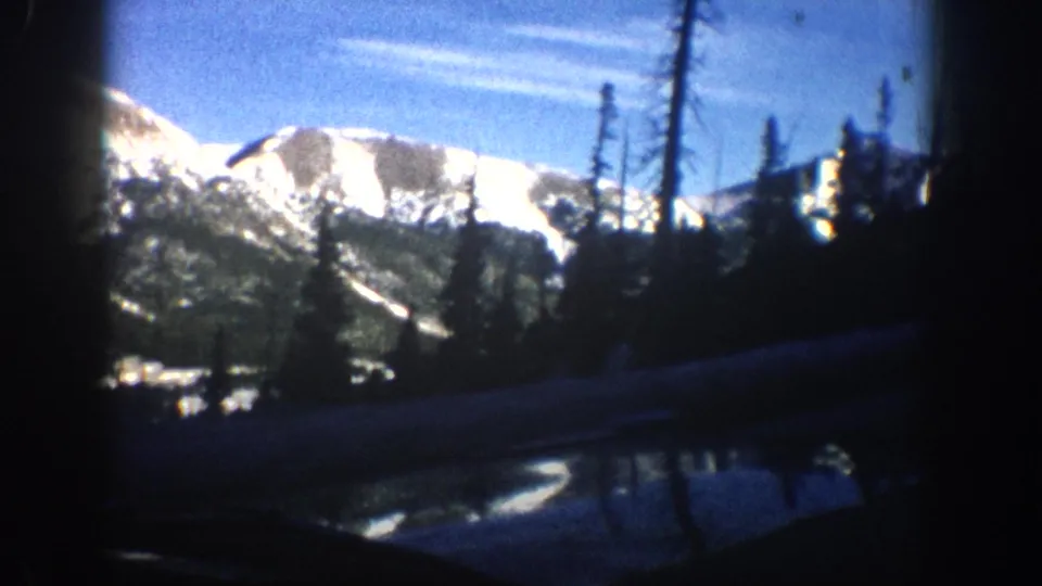 8mm home-movie frame showing broad snow-capped peaks above pine treeline and a wet mountain road surface, Aspen, Colorado area, circa 1961.