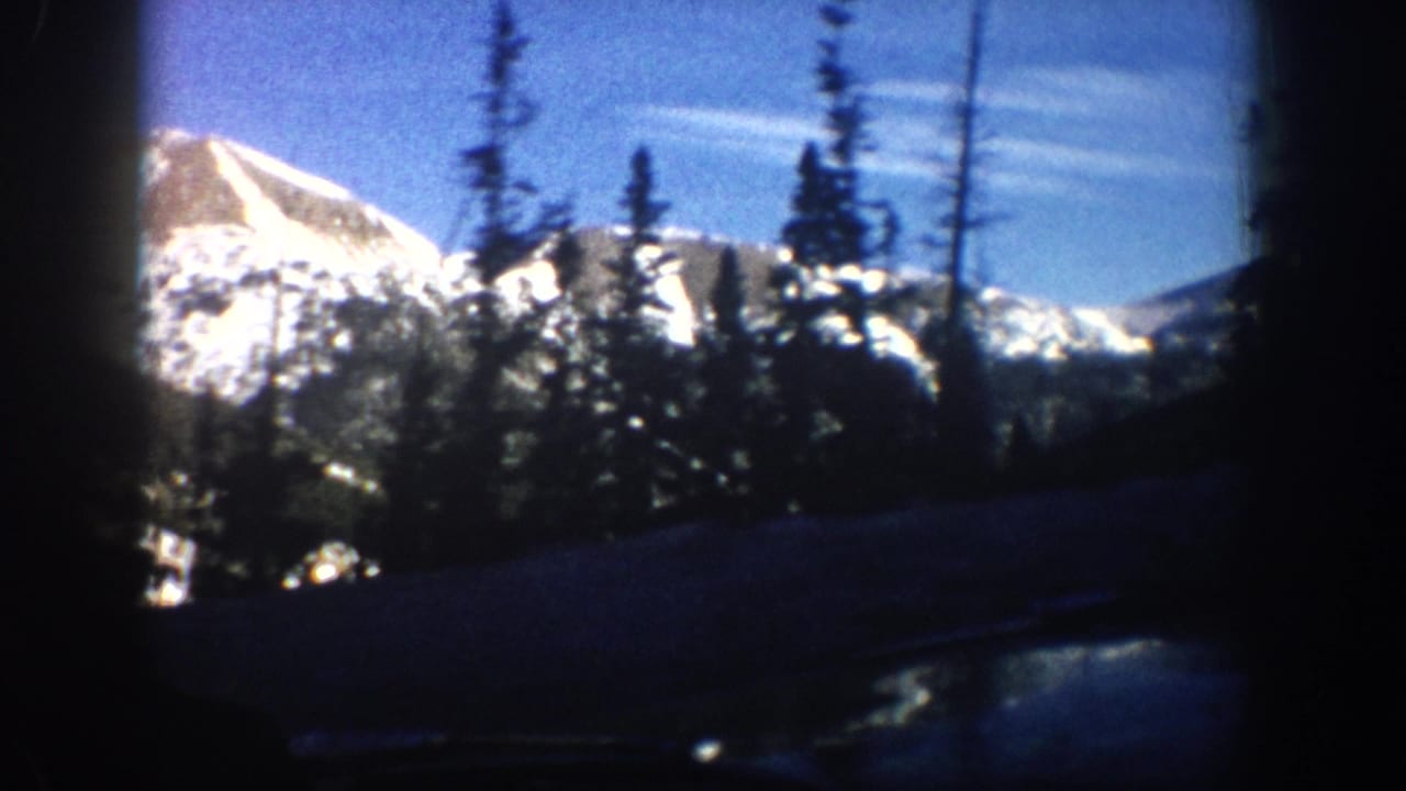8mm amateur footage from 1961 showing a snow-covered Rocky Mountain peak and dark evergreen trees framed by a car window edge, Aspen area, Colorado.