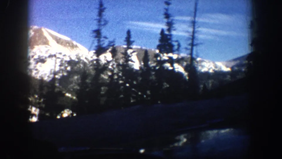 8mm amateur footage from 1961 showing a snow-covered Rocky Mountain peak and dark evergreen trees framed by a car window edge, Aspen area, Colorado.