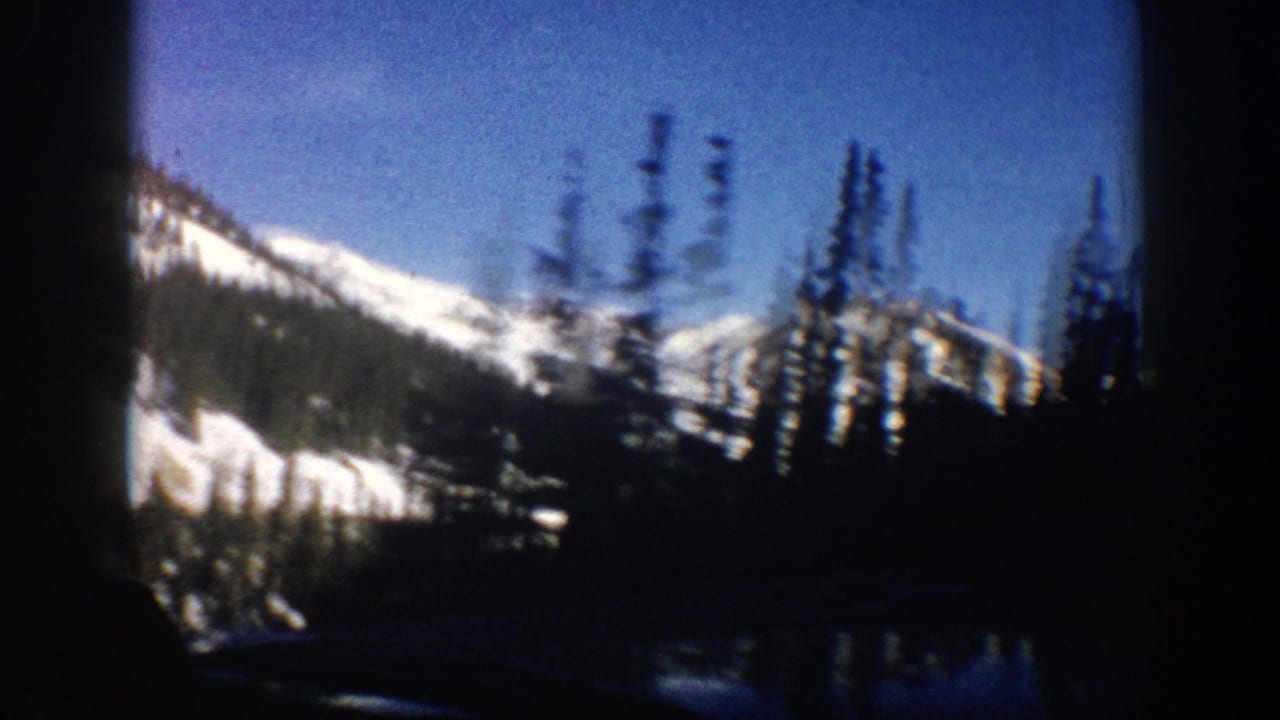 Broad snow-covered mountain slope and dark evergreen trees seen through a car window, 8mm film.