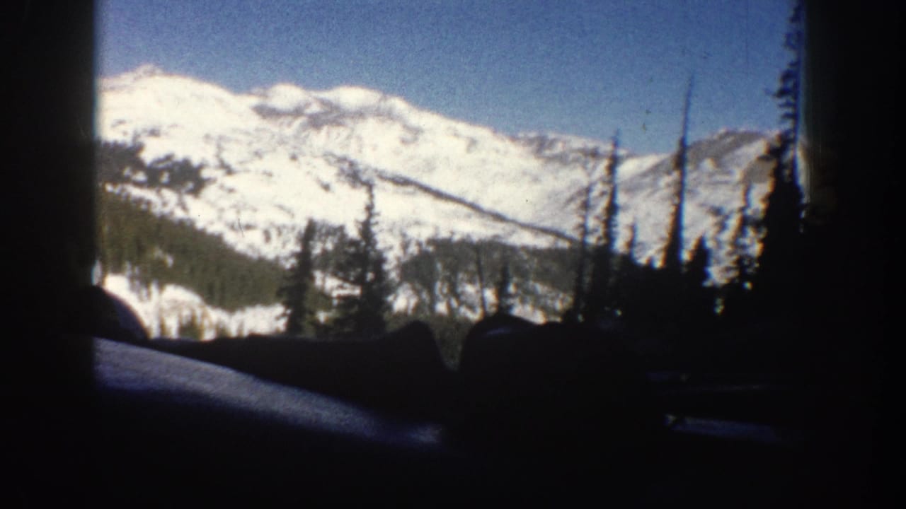 8mm home movie frame from Aspen, Colorado, 1961, showing a car-window view of snow-covered Rocky Mountain peaks with conifer trees in the foreground.